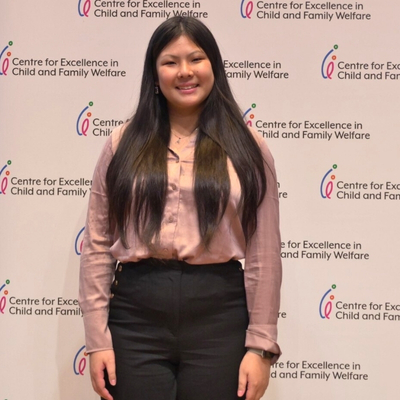 A young East Asian woman with long black hair, professionally dressed in a pink long-sleeved blouse and grey slacks, standing in front of a wall covered in the logo for the Centre for Excellence in Child and Family Welfare. She is smiling as has her hands down by her sides.