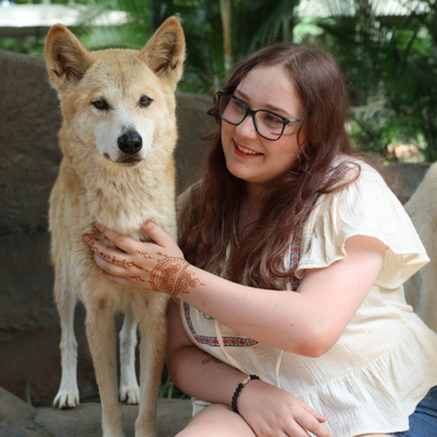 Photograph of a young woman with fair skin and long, brown hair wearing green glasses and a light summer blouse. She is sitting next to a white dog and smiling as she pats its chest. On her left hand, she has a henna tattoo.