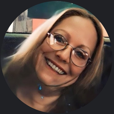 Heavily filtered photograph of a woman with straight blond hair and wire rimmed glasses smiling widely for the camera, leaning into frame from the left.