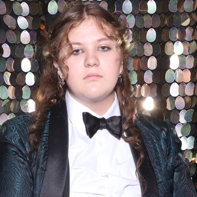 Photograph of a young woman with fair skin, blue eyes and long, curled, light brown hair. She is wearing a tuxedo with a blue lined pattern, silver earrings and eyeliner. Her expression is serious. Behind her is a wall of shiny event decorations.