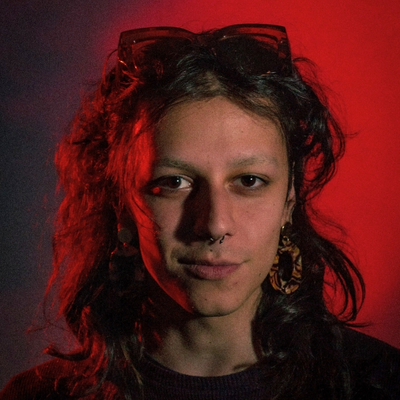 Photograph of person with olive skin, artfully messy shoulder-length hair, and brown eyes. They are wearing a black shirt, dangling earrings, a nose ring, and big plastic-framed glasses on their head. They are smirking slightly for the camera. The lighting behind them is red and moody.