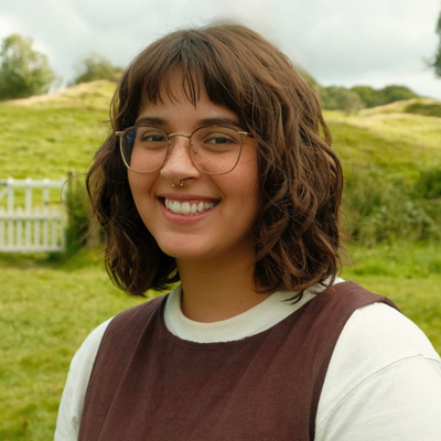 Photo of a young person with shoulder-length, wavy brown hair and olive skin smiling for the camera. They have a nose piercing and wire-rimmed glasses. They are wearing a casual burgundy vest over a white shirt. They are standing outside with rolling green fields and a white picket fence in the background.
