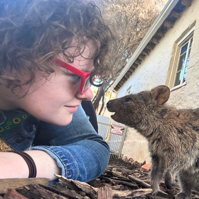 A young, fair-skinned woman with short, curly brown hair and red glasses, wearing a denim jacket and a hidden disabilities sunflower lanyard. She is down on the ground, nose to nose with a quokka. They are on tan bark next to an old house with a pale yellow render over a brick wall.