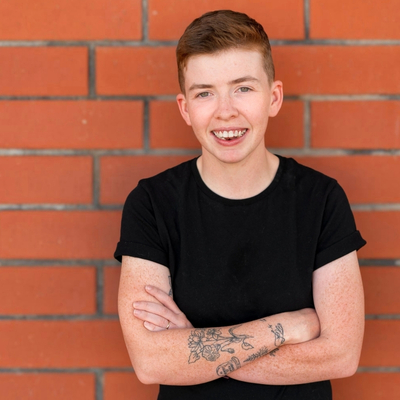 Photograph of a young, fair-skinned person with short cropped, light brown hair and blue eyes, wearing a black t-shirt. Their arms are crossed over their chest. Their right arm has multiple tattoos, with a tattoo of a bunch of flowers most visible. They are leaning back against a red brick wall and smiling for the camera.