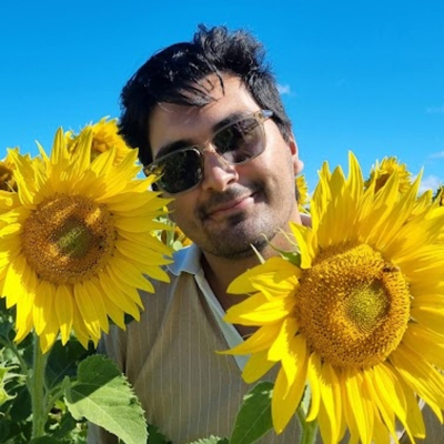 A smiling young man with brown skin, short black hair and a five o'clock shadow, wearing sunglasses and a pin striped shirt. He is outside in a field of giant sunflowers with a clear blue sky behind him.