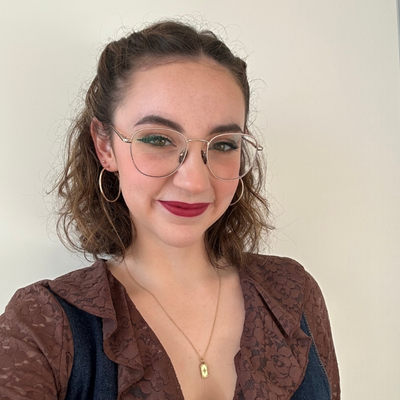 Selfie of a young, fair-skinned woman with wire-rimmed glasses and shoulder-length, curly brown hair, wearing professional make-up, hoop earrings, a gold-chain necklace and a lacy brown, floral blouse. She is smiling for the camera.