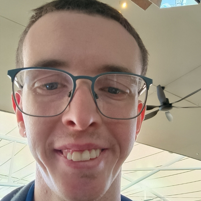 Photograph of a young man with fair skin, short cropped light brown hair and a buck-toothed smile wearing wire rimmed glasses. He is looking down at the camera. Behind him, we can see a ceiling fan and white rafters.