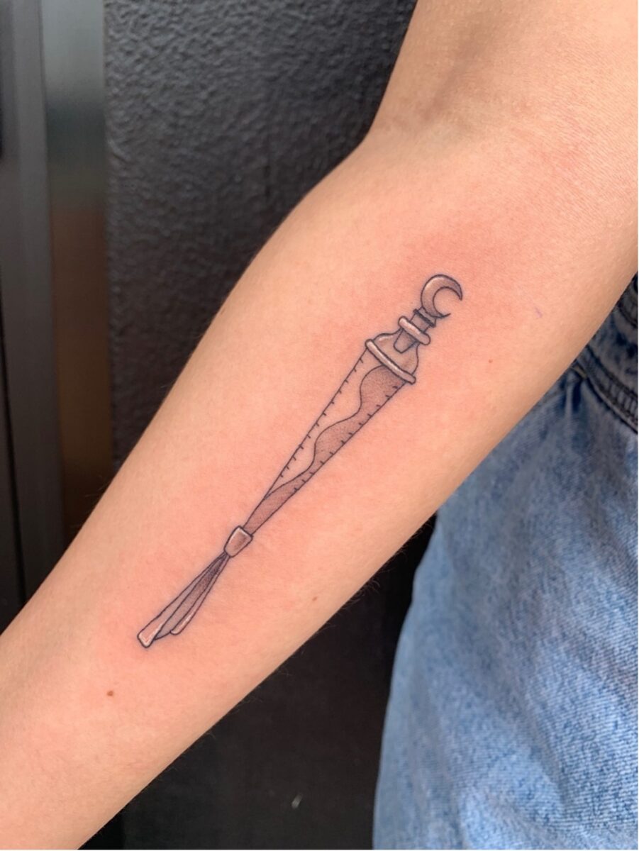 A black linework tattoo on Chloe's forearm of Katara's amulet of spirit water from Avatar the Last Airbender. The design shows a slender pendant with wave-like patterns, a small crescent moon at the top, and a tassel at the bottom.
