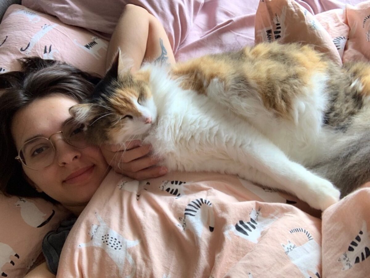 A selfie of a young adult with olive skin, shoulder-length brown hair and wire-rimmed glasses. They are lying in bed with their tortoiseshell cat draped over them and bed covers pulled up to their chin. The bed cover and pillow cases are sand-coloured with a pattern of black and white cats in various poses.