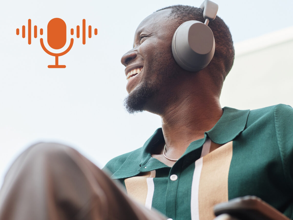 Photograph of a young, black man with a big smile and a goatee wearing a green and gold polo shirt and large headphones. The photo is taken from below and the arm of the man's wheelchair is just visible at the bottom of the frame. The sky and a white building can be seen behind him. An orange graphic of a microphone is superimposed over the photo in the top-left corner.