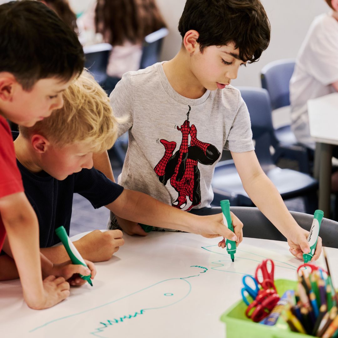 A photo of three boys using green markers to draw a snake and other pictures on a large sheet of white paper. They are in a classroom. The boy on the right has dark hair and is wearing a T-shirt that features the Marvel character Spider-Man. The boy in the middle has short blonde hair and is wearing a black top. The boy on the left has dark hair and is wearing a red t-shirt.