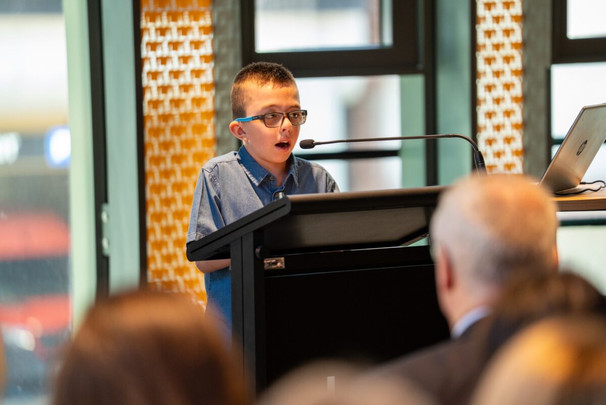 A photo of Jerzy, a 10-year-old boy swith short dark blond hair wearing a blue button-up top and glasses. He is standing at a lectern and speaking into a microphone in front of a crowd.