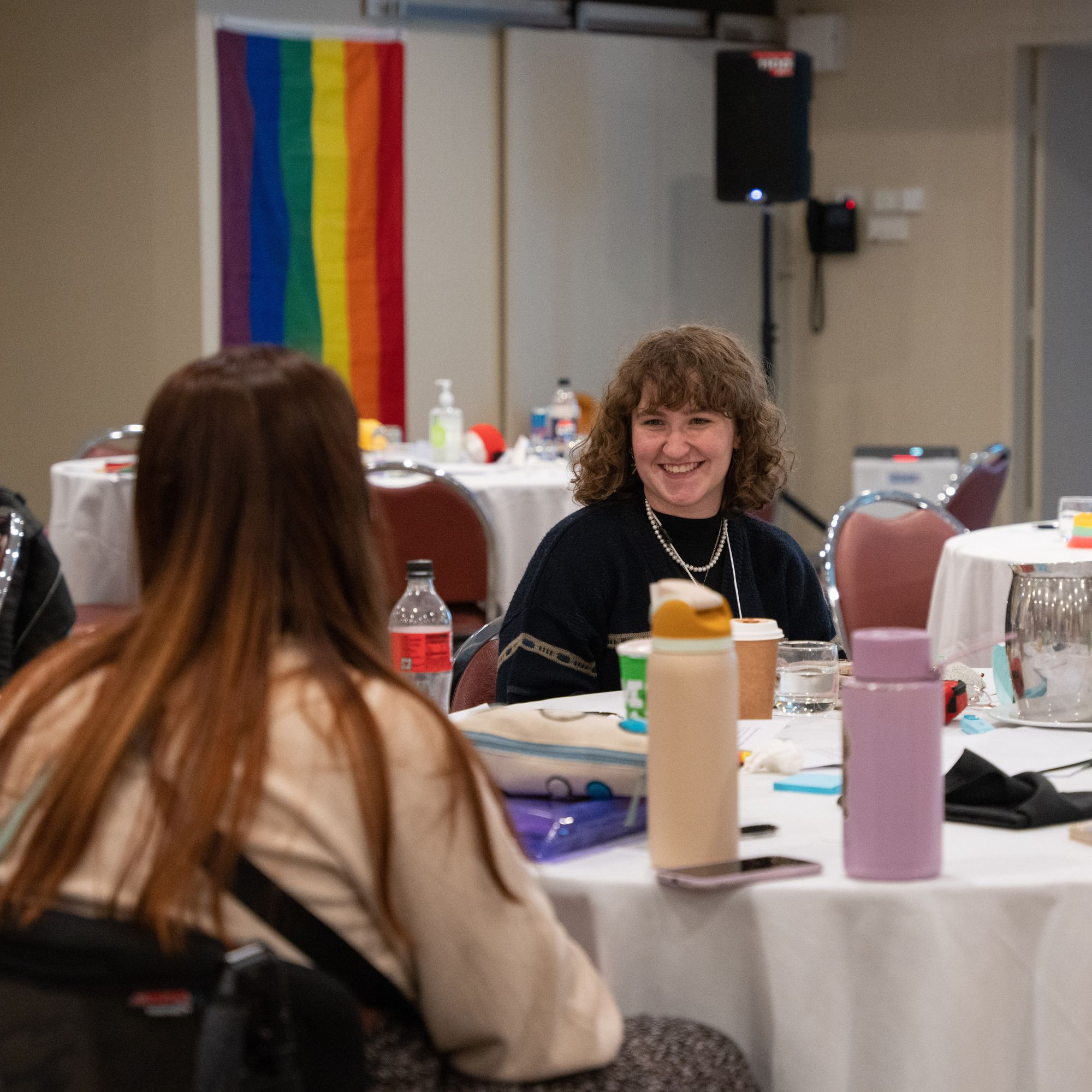 A photo of two young people sitting at table in a brightly lit conference room. The table is covered in bottles and writing utensils. A rainbow LGBTQIA+ flag can be seen hanging on a wall in the background.