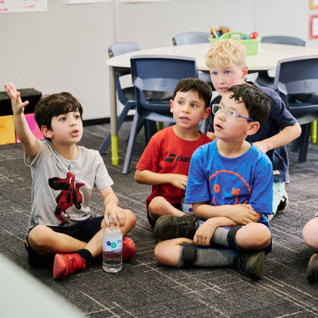 A photo of four young boys sitting on the floor of a classroom. The boy on the left is raising his hand.