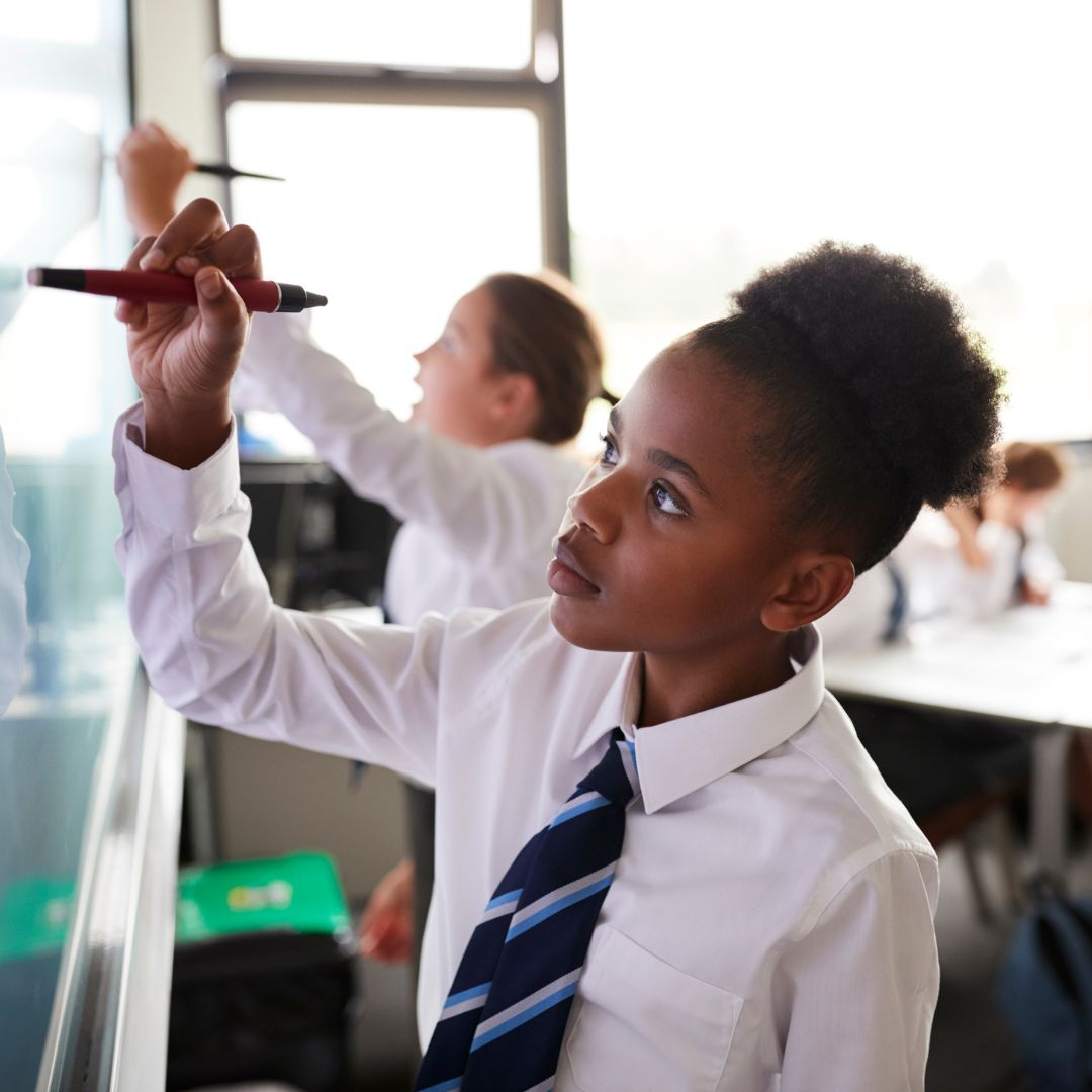 A photo of a teenage girl wearing a school uniform that includes a crisp white shirt and a striped navy tie. She is using a marker to write on a white board.