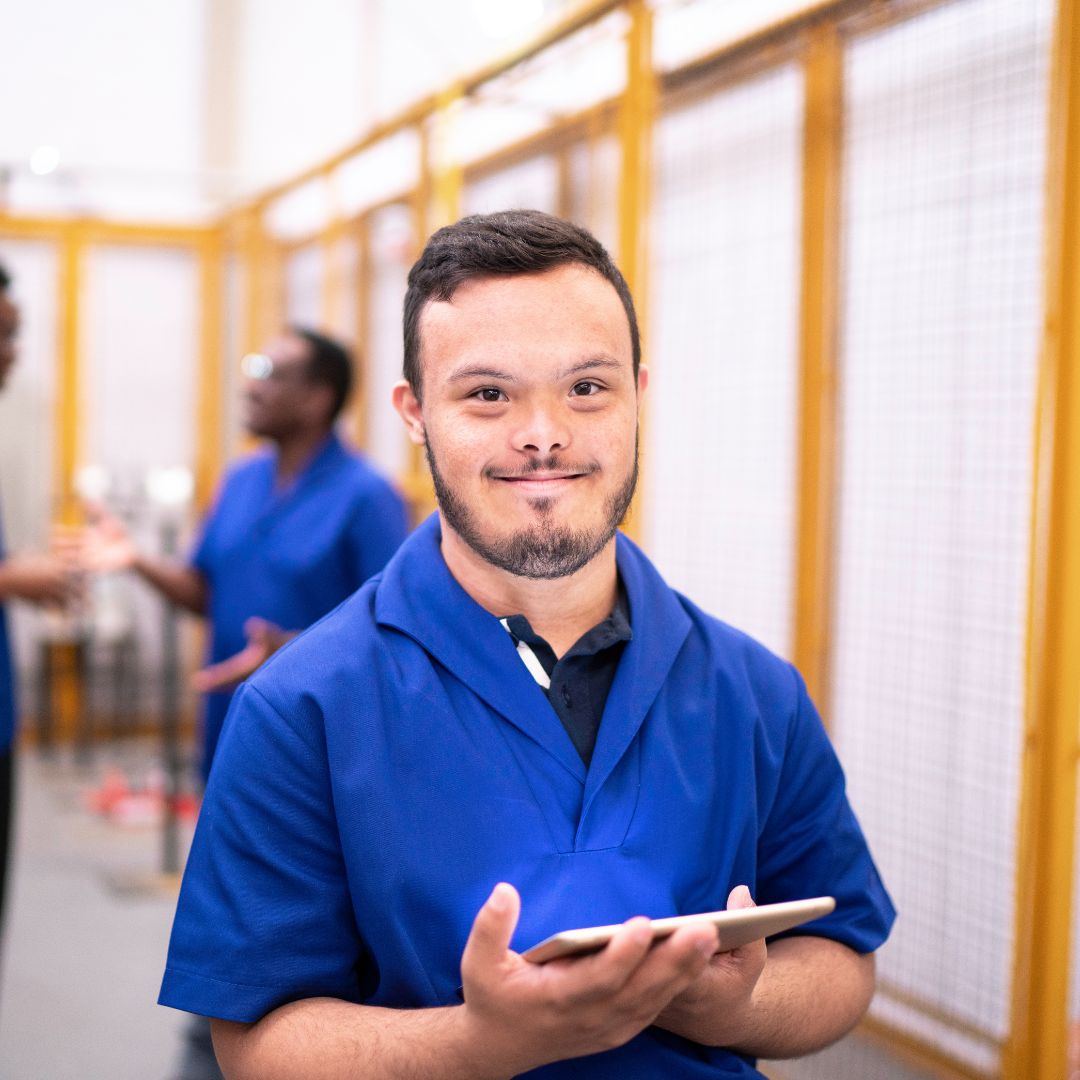 A photo of a young man at a worksite. He has short dark hair, is wearing a blue uniform, and is holding a tablet while smiling.