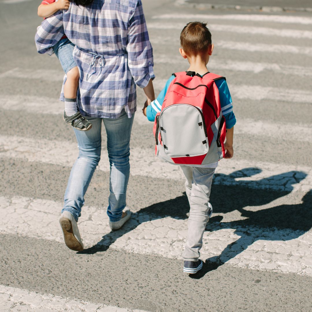 A photo of a child wearing a red backpack holding the hand of an adult as they cross a crosswalk. The adult is holding a younger child in their left arm.