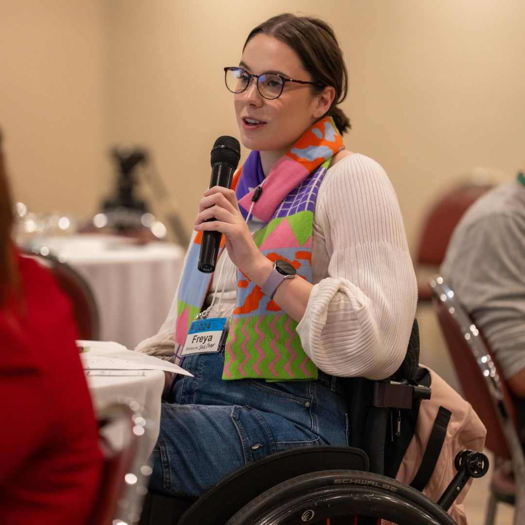 A photo of a young woman with mid-length brown hair and glasses speaking into a microphone at an event. She is wearing a bright coloured vest and is using a wheelchair.