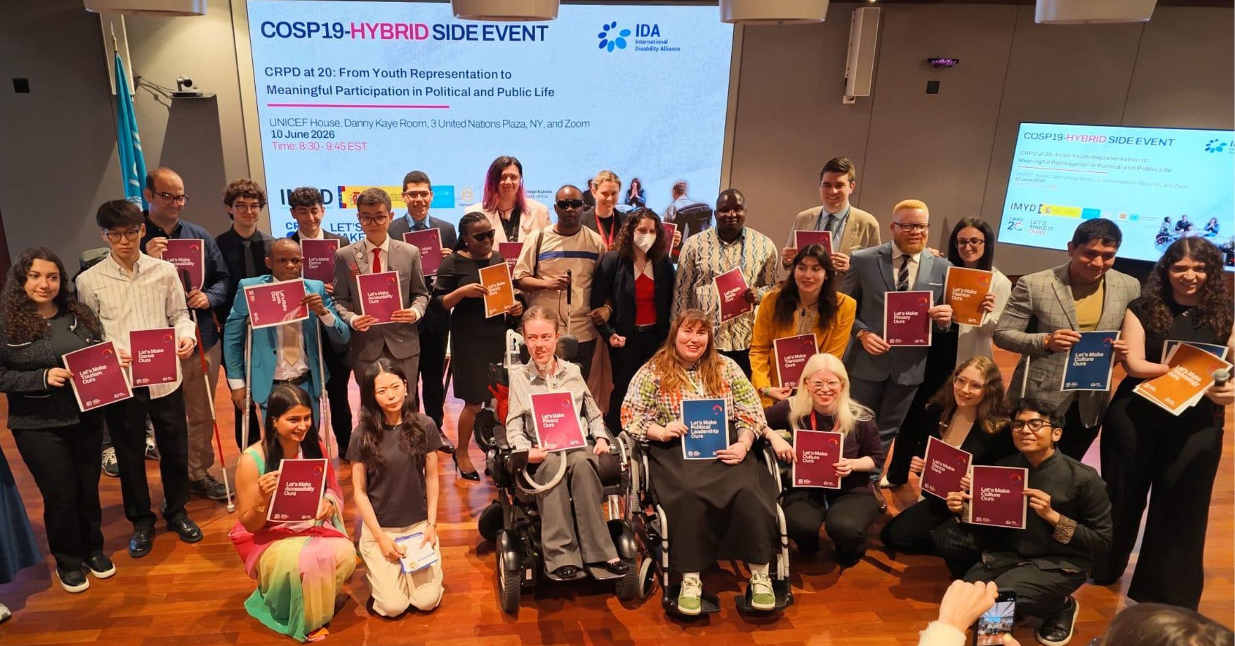 A photo of a large group of young people with disability smiling and posing for a photo in a convention space. A screen behind the group features text that reads: "COSP19-Hybrid Side Event. CRPD at 20: From Youth Representation to Meaningful Participation in Political and Public Life."
