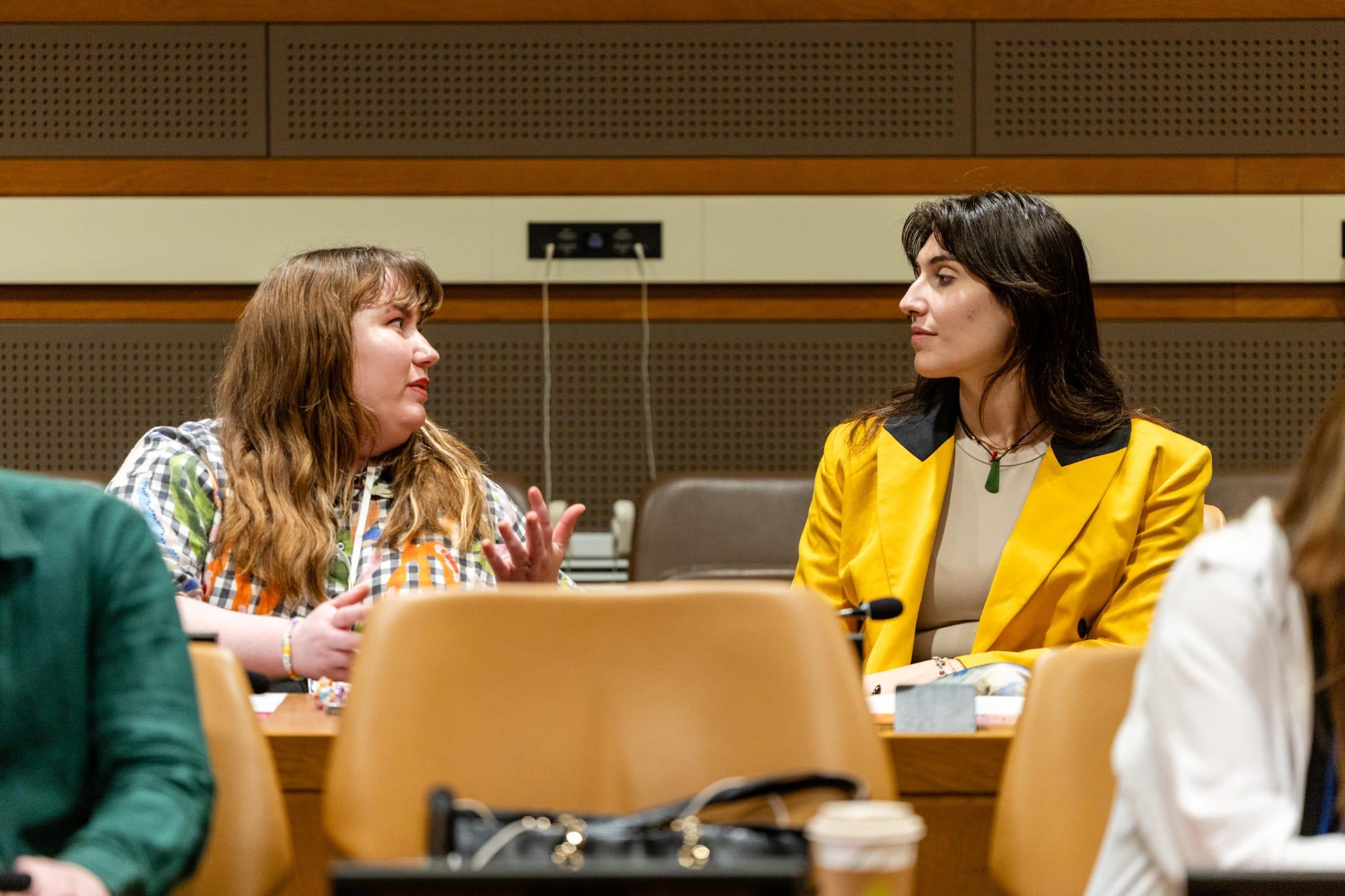 A photo of two young women in conversation at a table in a hall at the United Nations. The woman on the left has long orange hair and is gesturing as she speaks. The woman on the right has long dark brown hair and is wearing a bright yellow blazer.