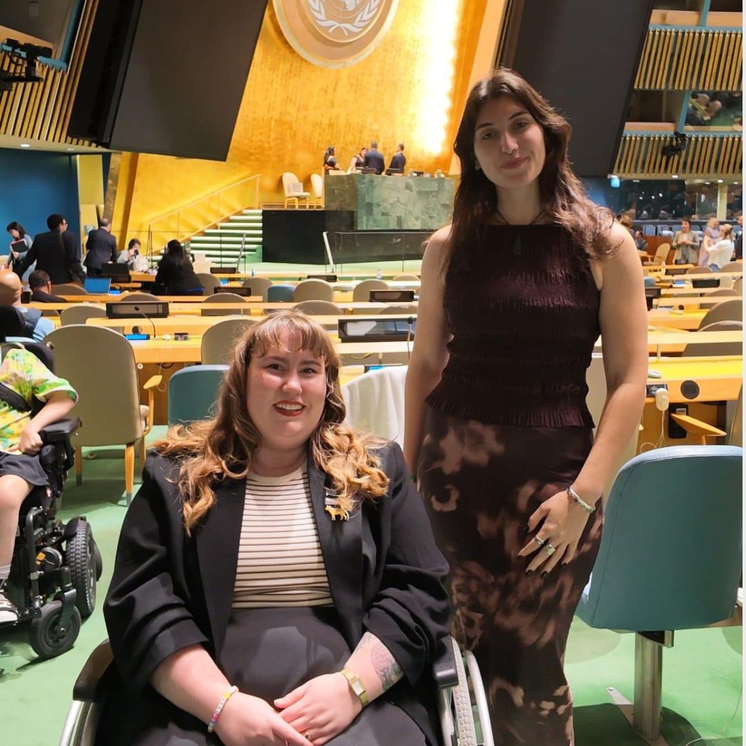 A photo of two young women side by side in the General Assembly hall at the United Nations. The woman on the left has long orange hairing and is wearing a black blazer and using a wheelchair. The woman on the right is wearing a dark dress and has long brown hair. Both are smiling. Behind them, the UN crest is mounted on a brightly lit wall.