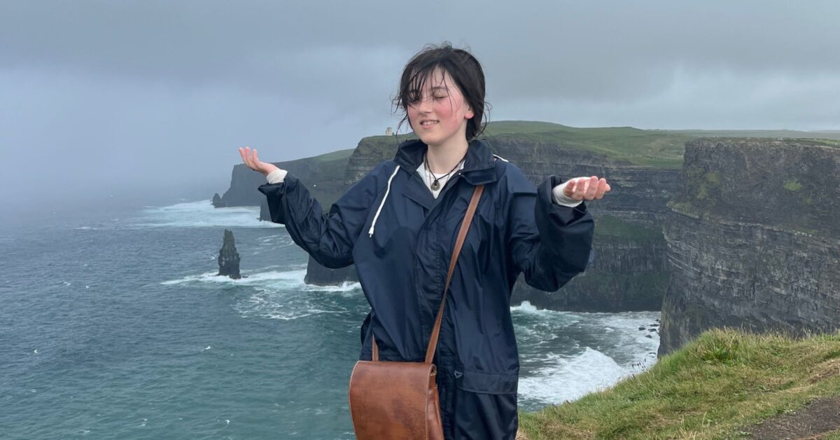 A photo of Faith, a young caucasian woman standing on a cliffside overlooking the ocean on an overcast day. They are wearing a blue raincoat and holding their hands out.