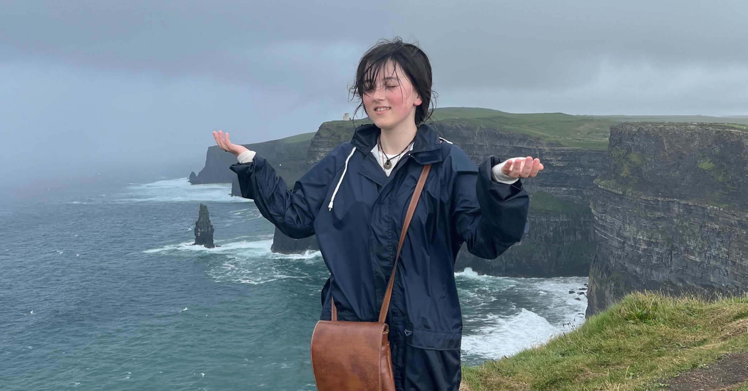 A photo of Faith, a young caucasian woman standing on a cliffside overlooking the ocean on an overcast day. They are wearing a blue raincoat and holding their hands out.
